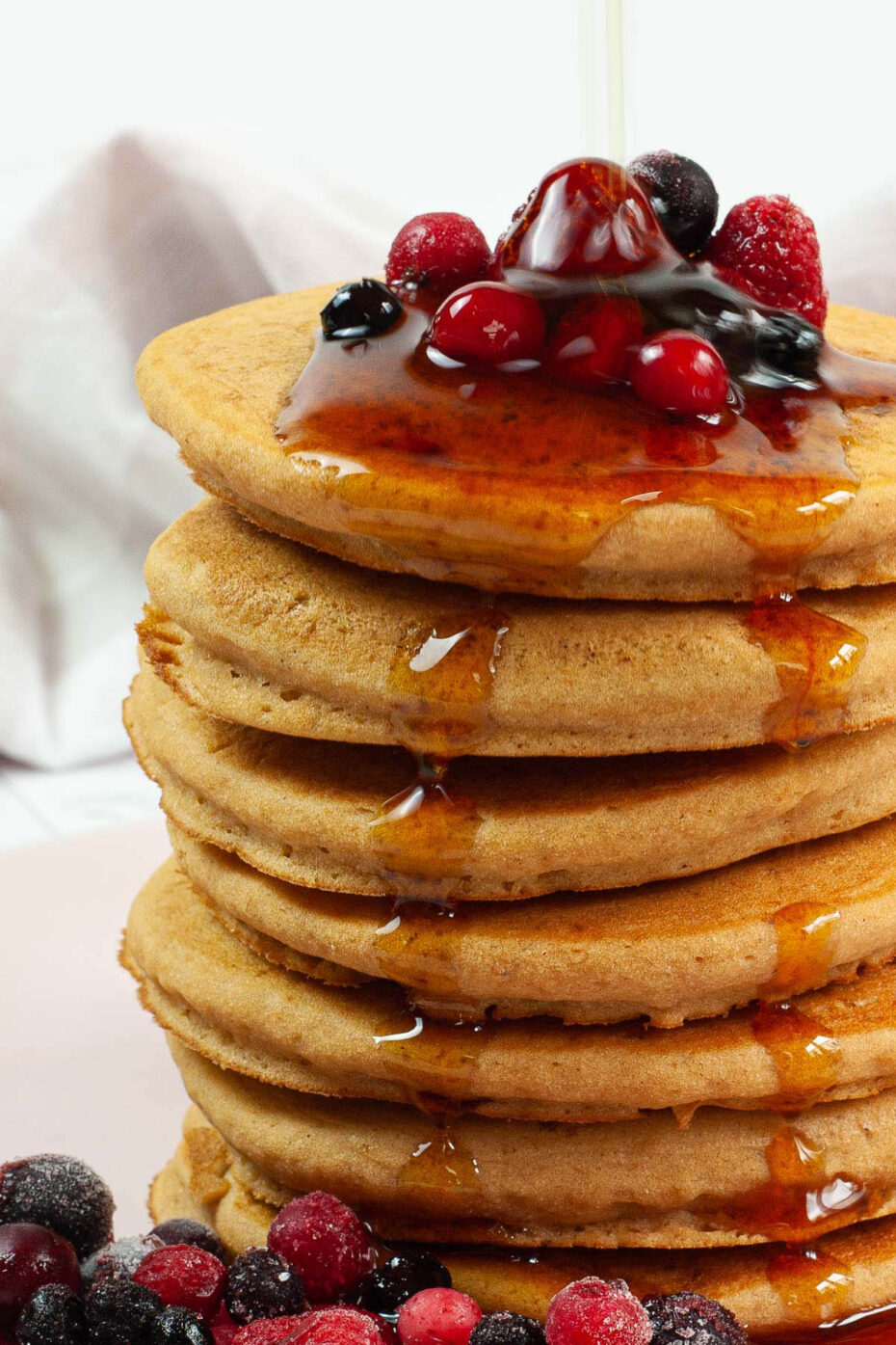 Pink plate with a stack of pancakes topped with black and red berries and maple syrup is pouring from the top along the sides.