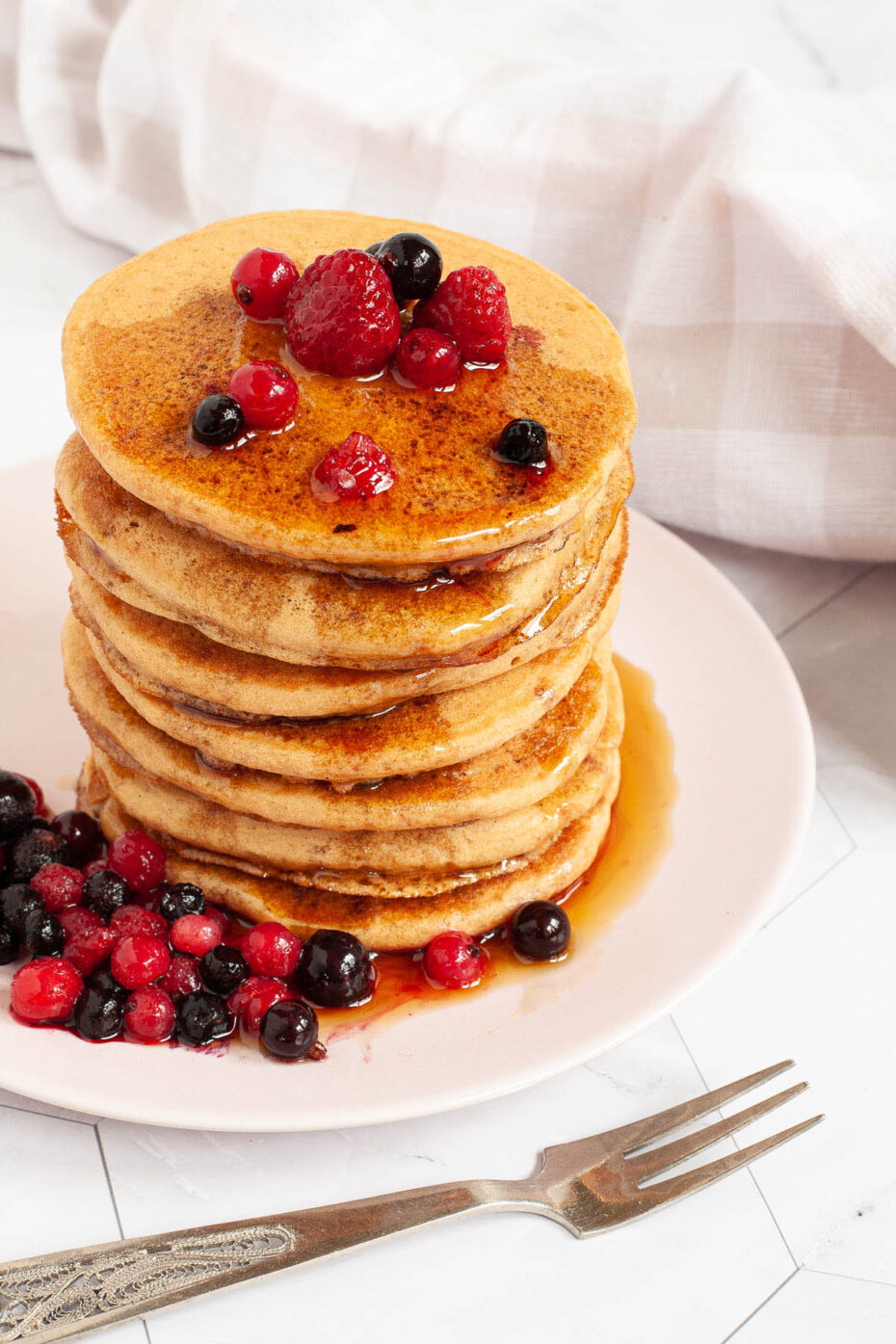 Pink plate with a stack of pancakes topped with black and red berries and maple syrup