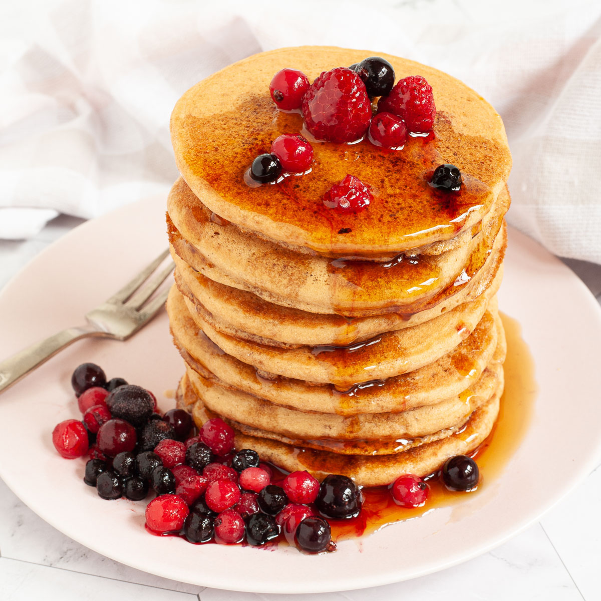 Pink plate with a stack of pancakes topped with black and red berries and maple syrup