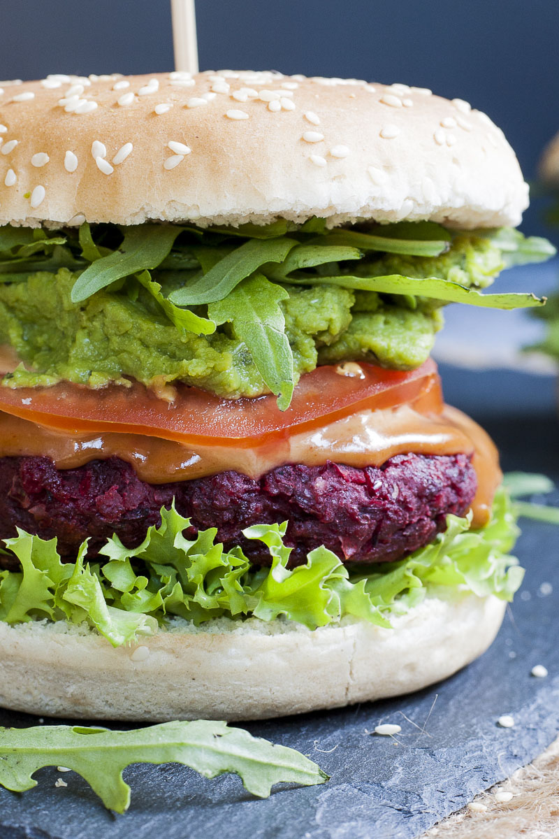 Close-up of a beet burger with layers like burger bun, lettuce, purple patty, tomato slice, green puree, arugula and bun on the top