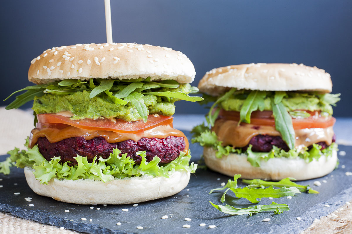 Close-up of a beet burger with layers like burger bun, lettuce, purple patty, tomato slice, green puree, arugula and bun on the top