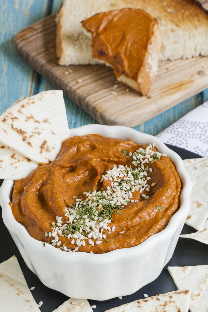 Orange brownish cream in a white bowl sprinkled with green dried herbs and tiny white seeds. Toast with orange brownish cream in the back. Pita chips scattered around. 