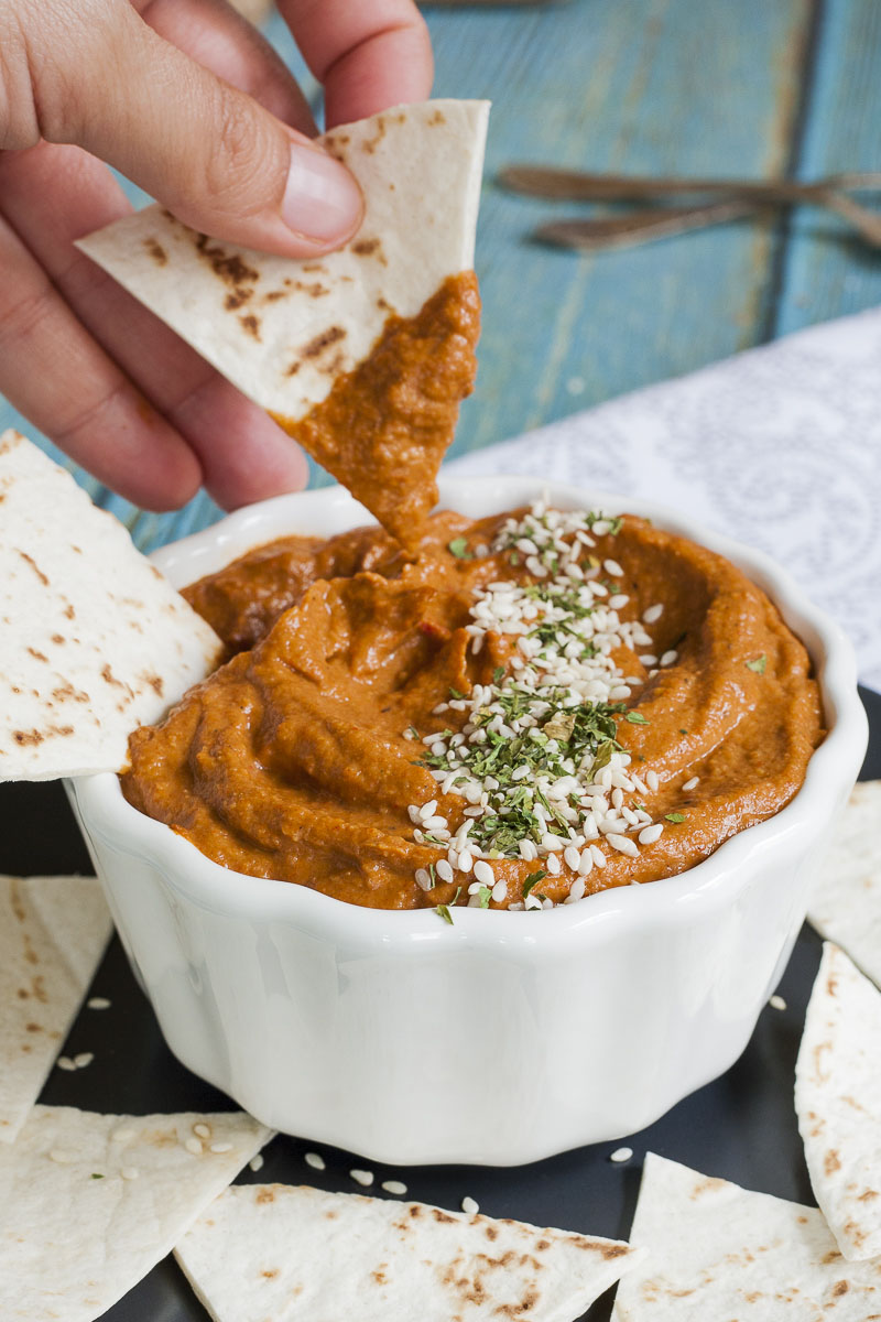 Orange brownish cream in a white bowl sprinkled with green dried herbs and tiny white seeds. A hand is dipping a pita triangle in it. 