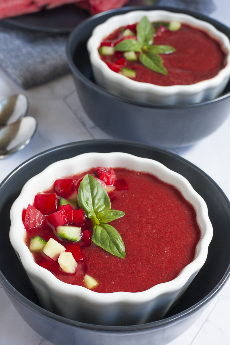 Red soup in 2 white and black bowls topped with basil leaves and diced red and green veggies.