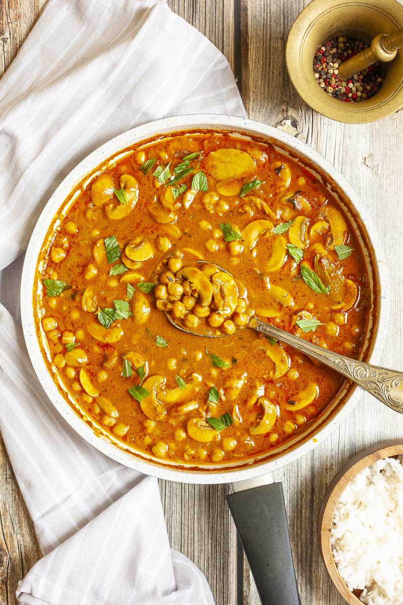 Large saucepan from above with brown-yellow liquid chickpeas and mushroom slices sprinkled with chopped green leaves