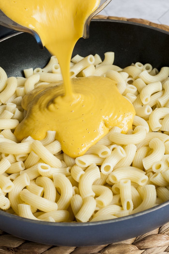 A frying pan is full of pasta. A hand is pouring a yellow sauce on it directly from a blender. 
