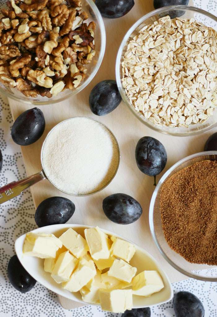 Small bowls with different ingredients to make plum crumble. One with butter cubes, one with rolled oats, one with walnut halves, one with brown sugar and one with flour. Plums are scattered around