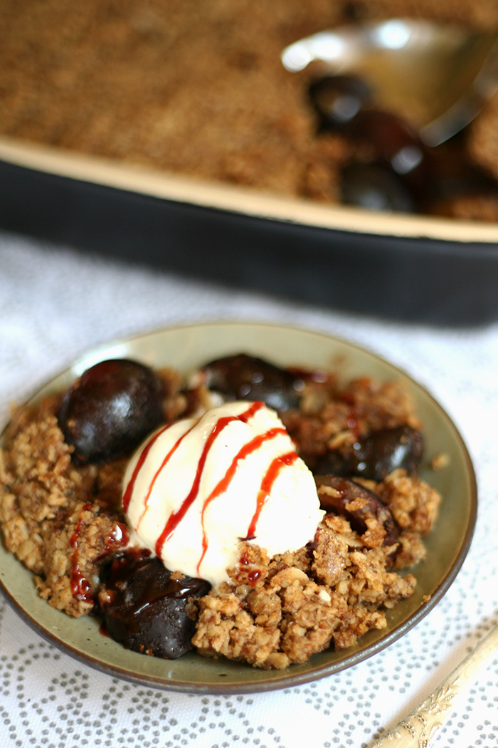 Small green plate with a serving of brown crumble, plum and a scoop of vanilla ice. The baking dish is in the background