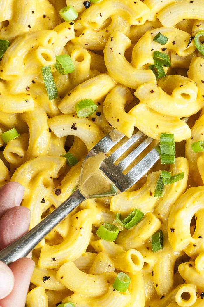 Pasta with yellow sauce, chili flakes and chopped spring onion. A hand is holding a fork and taking some. 