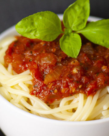White bowl is full of spaghetti with red chunky pasta sauce and a couple of basil leaves on top.