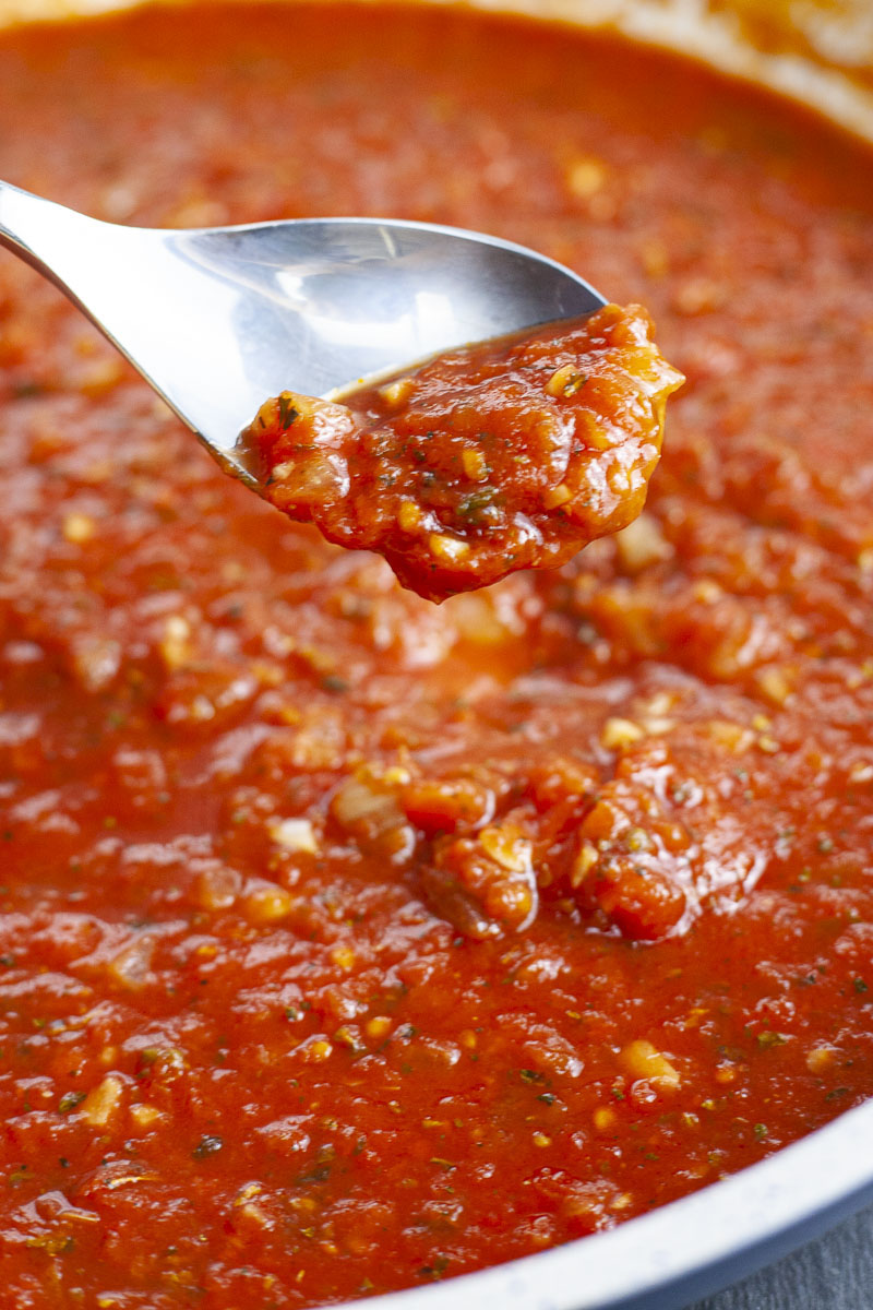 A large saucepan with lots of chunky red sauce with yellow and green bits and pieces ready to be served on pasta. A spoon is taking a small amount.