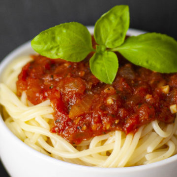 White bowl is full of spaghetti with red chunky pasta sauce and a couple of basil leaves on top.