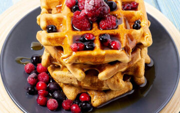 Black plate with a stack of waffles topped with mixed berries and maple syrup