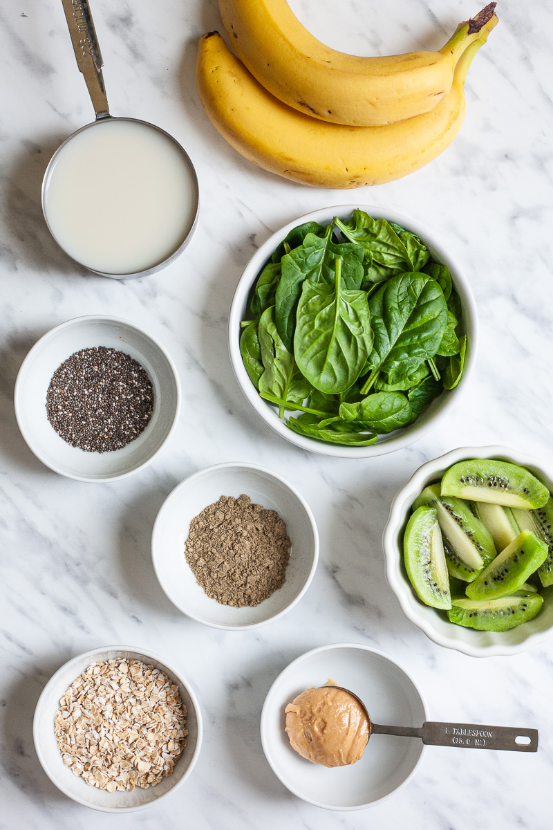 All ingredients in small bowl on a flat surface: milk, banana, spinach leaves, kiwi halves, oats, chia seeds, peanut butter