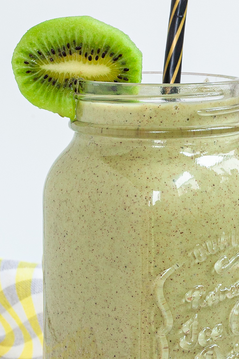 A large smoothie glass is filled with green liquid decorated with a kiwi slices and a black and gold straw. You can see 2 bananas and a couple of spinach leaves around it.