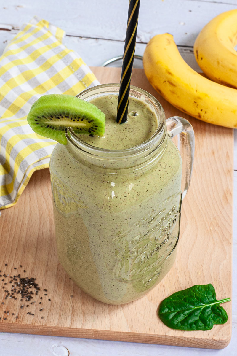 A large smoothie glass is filled with green liquid decorated with a kiwi slices and a black and gold straw. You can see 2 bananas and a couple of spinach leaves around it.