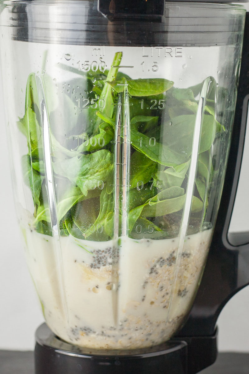 A blender is filled with different ingredients. At the bottom milk with oats and chia seeds and spinach leaves on the top.