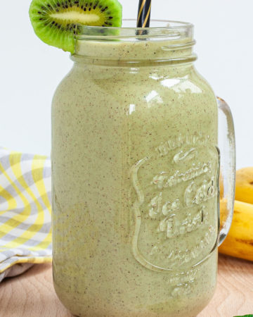 A large smoothie glass is filled with green liquid decorated with a kiwi slices and a black and gold straw. You can see 2 bananas and a couple of spinach leaves around it.