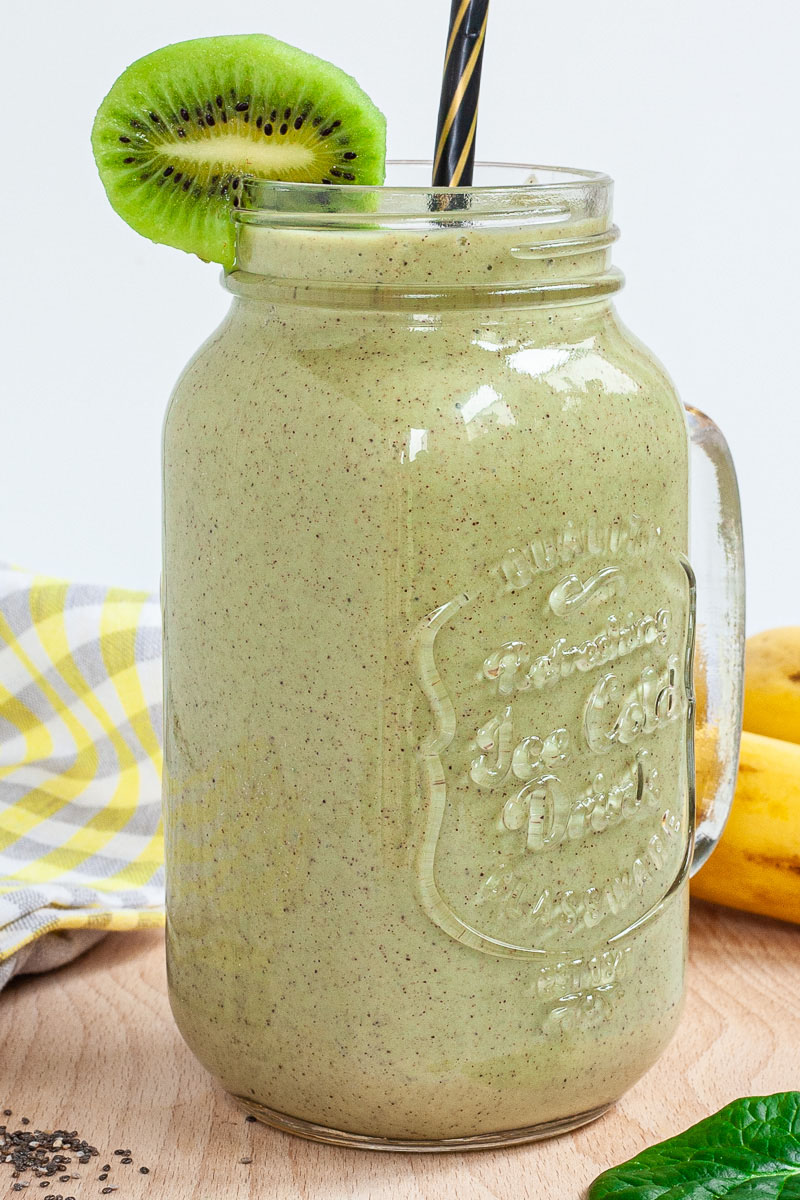 A large smoothie glass is filled with green liquid decorated with a kiwi slices and a black and gold straw. You can see 2 bananas and a couple of spinach leaves around it.