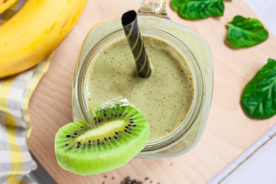 A large smoothie glass is filled with green liquid decorated with a kiwi slices and a black and gold straw. You can see 2 bananas and a couple of spinach leaves around it.