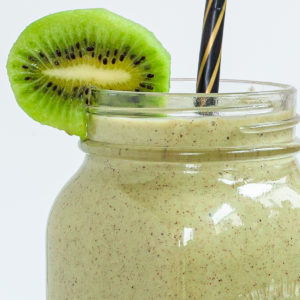 A large smoothie glass is filled with green liquid decorated with a kiwi slices and a black and gold straw. You can see 2 bananas and a couple of spinach leaves around it.