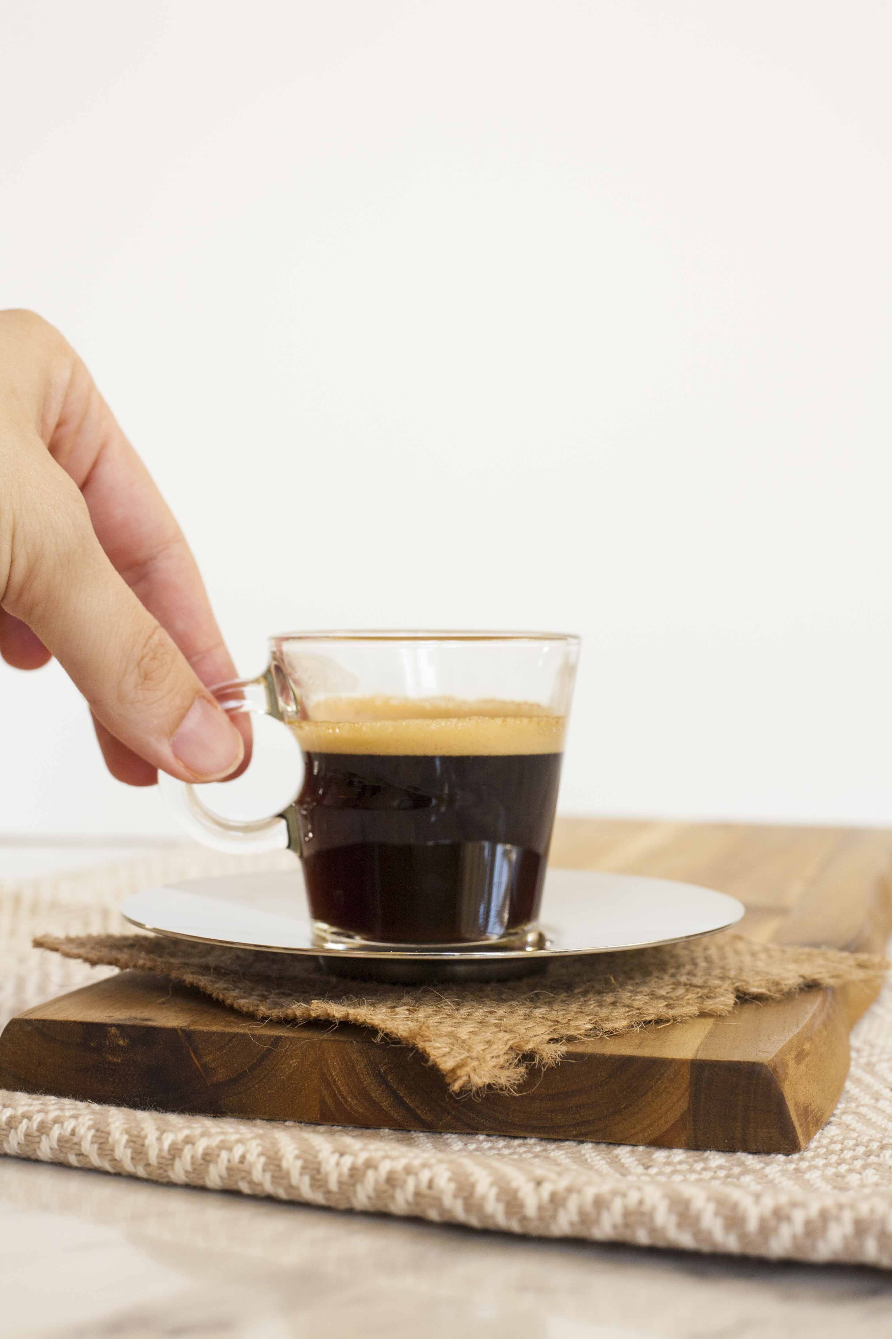A small espresso glass with dark brown coffee. A hand is holding the glass by its ear. It is placed on a silver plate. 