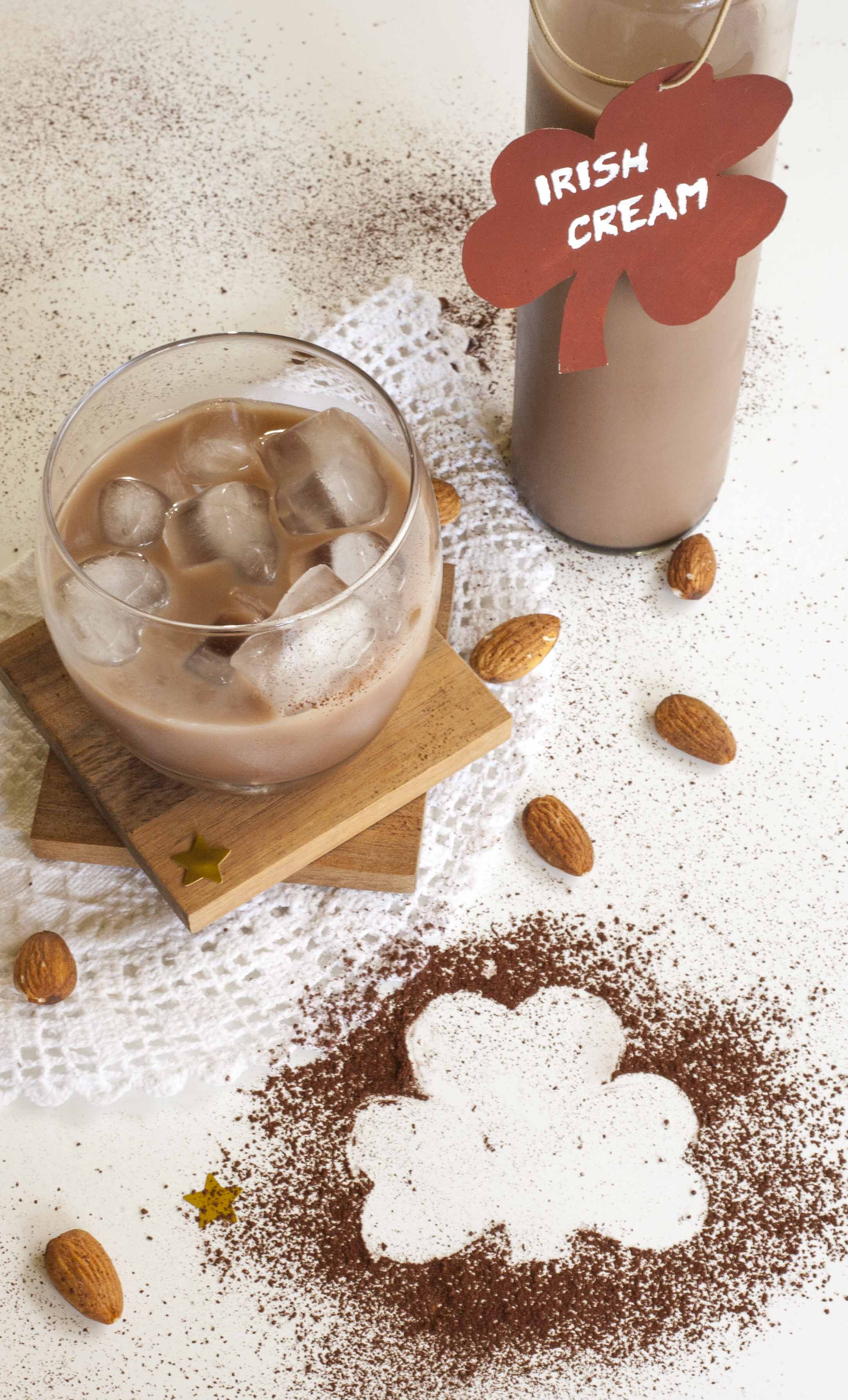 A whiskey glass with brown creamy drink and heart-shaped ice. A tall bottle full of brown creamy drink is next to it with a label with a name, Irish cream. Almonds and cocoa powder are around them. 