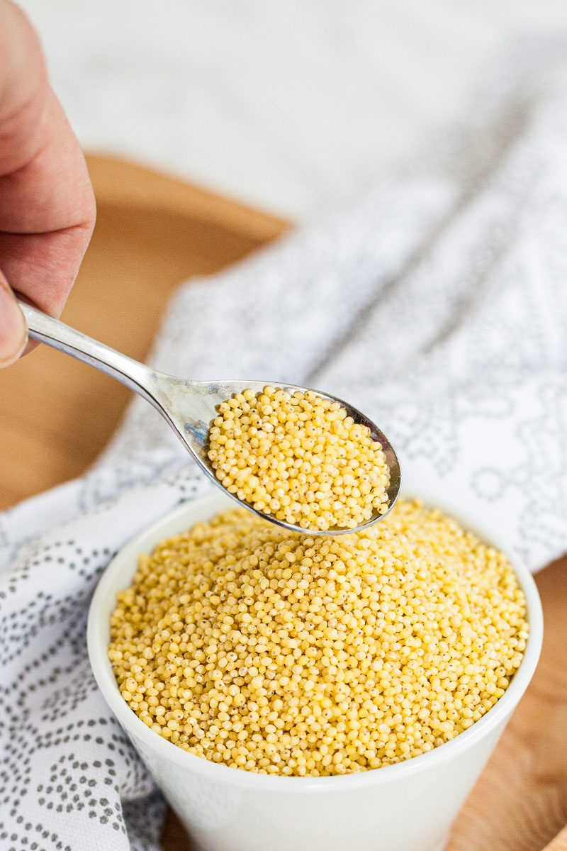 Small white bowl full of yellow grains. A hand holding a spoon is taking small from the top.