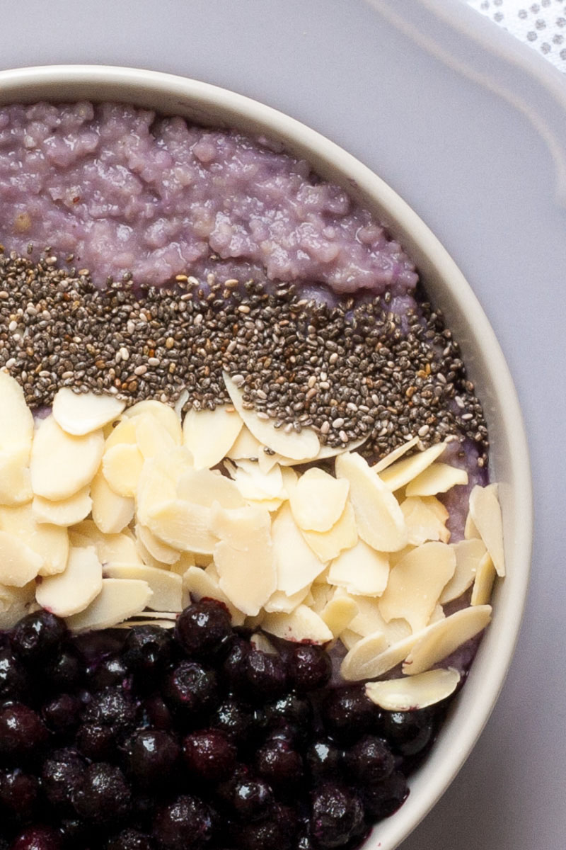 A white bowl on a light purple plate full of purple porridge topped with small grey seeds, almond slices and lots of purple blueberries