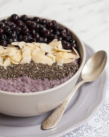 A white bowl on a light purple plate full of purple porridge topped with small grey seeds, almond slices and lots of purple blueberries