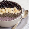 A white bowl on a light purple plate full of purple porridge topped with small grey seeds, almond slices and lots of purple blueberries