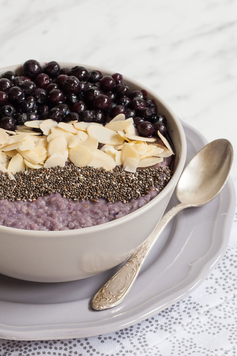 A white bowl on a light purple plate full of purple porridge topped with small grey seeds, almond slices and lots of purple blueberries