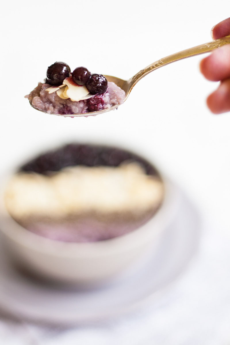 A spoon with purple porrdige and blueberries on top. A white bowl on a light purple plate full of purple porridge topped with small grey seeds, almond slices and lots of purple blueberries in the background