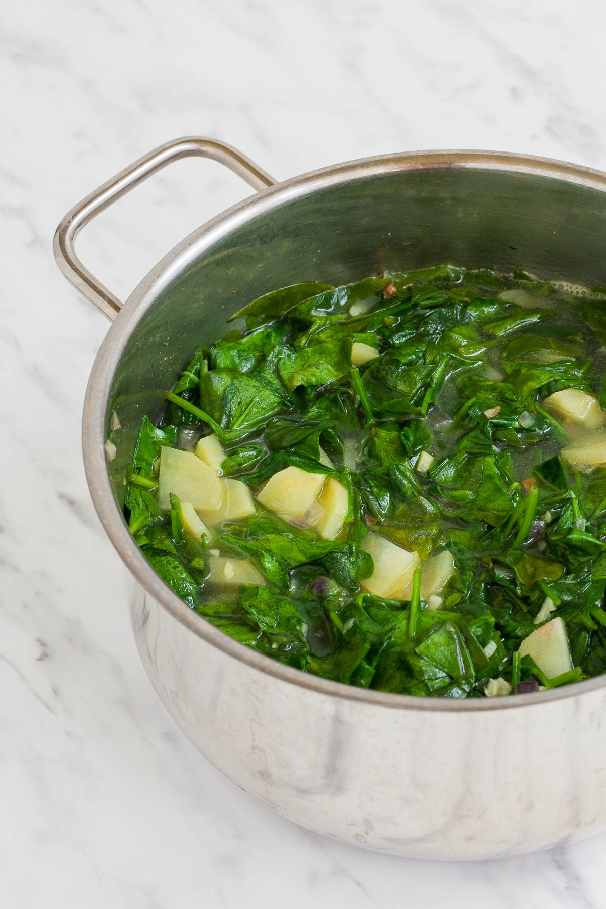 Silver stockpot with spinach soup where you can see the ingredients chopped potatoes, onion, garlic pieces and wilted spinach leaves