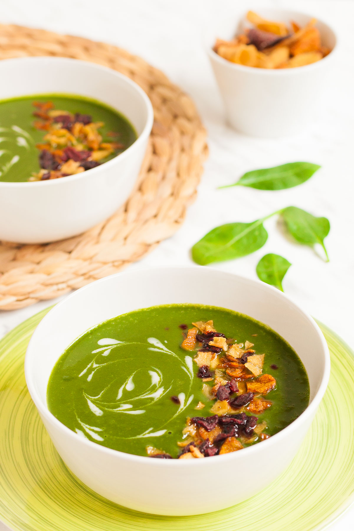 2 bowls of green soup sprinkled with vegetable crisps and white swirls.