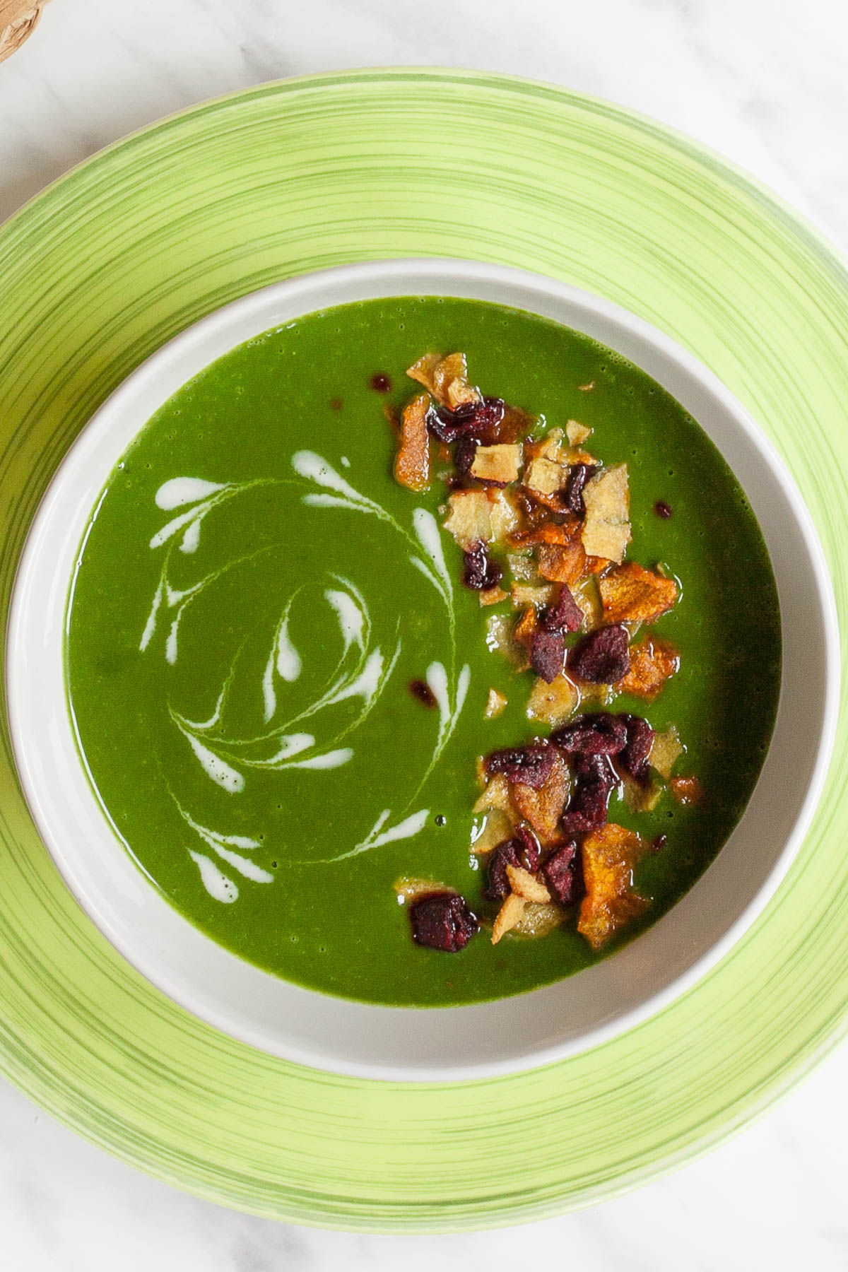 A bowl of green soup sprinkled with vegetable crisps and white swirls.