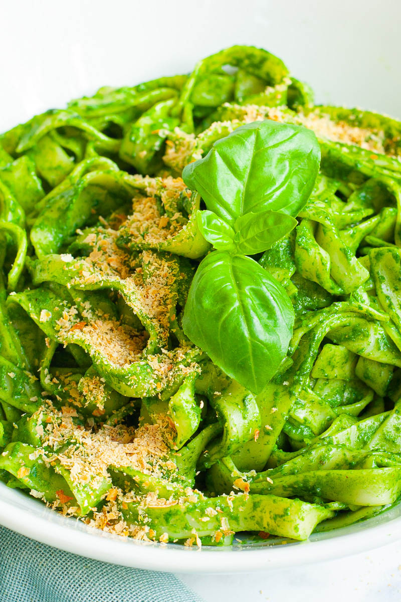 White bowl with fettuccine pasta covered in green sauces sprinkled with yellow flakes and a pair of basil leaves.