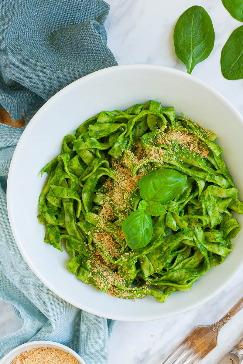 White bowl from above with fettuccine pasta covered in green sauces sprinkled with yellow flakes and a pair of basil leaves. A couple of leaves are scattered around. There is a small white bowl with more yellow flakes.