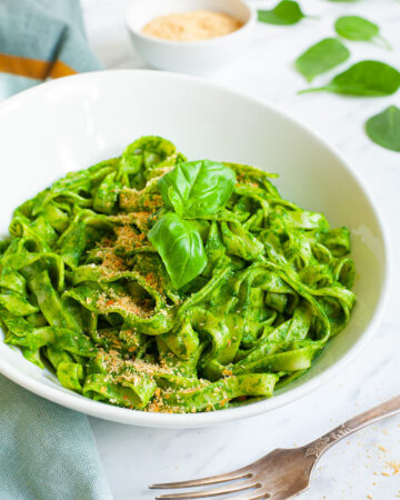 White bowl with fettuccine pasta covered in green sauces sprinkled with yellow flakes and a pair of basil leaves. A fork is next to the bowl.