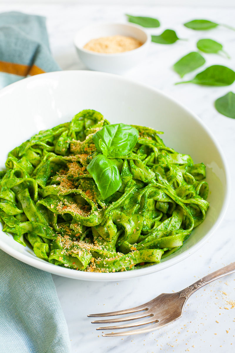 White bowl with fettuccine pasta covered in green sauces sprinkled with yellow flakes and a pair of basil leaves. A fork is next to the bowl.