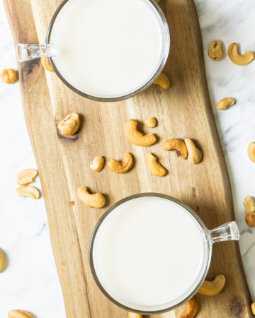 Two glass mugs on a wooden board shown from the top full of cashew milk. Cashew nuts sprinkled around.