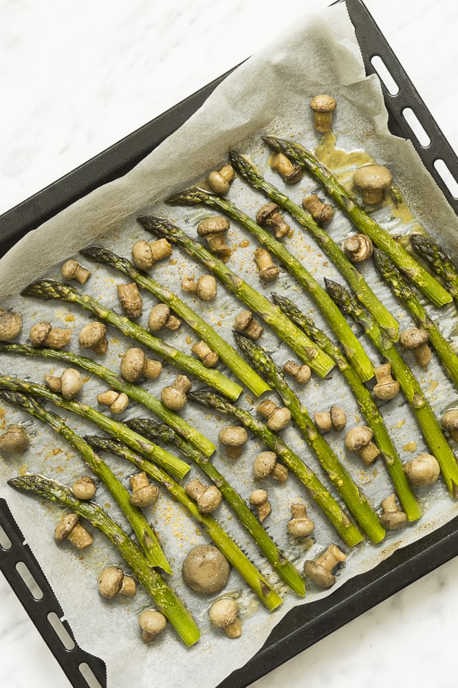 Black sheet pan full of roasted asparagus spears and small champion mushrooms on parchment paper