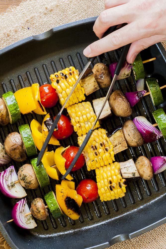 Close-up of grilled vegetable kabobs in a grill pan with tongs in hand