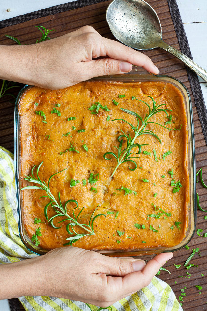 A glass casserole dish with layers of potato slices with an orange creamy sauce sprinkled with chopped parsley and rosemary. two hand is holding the sides.