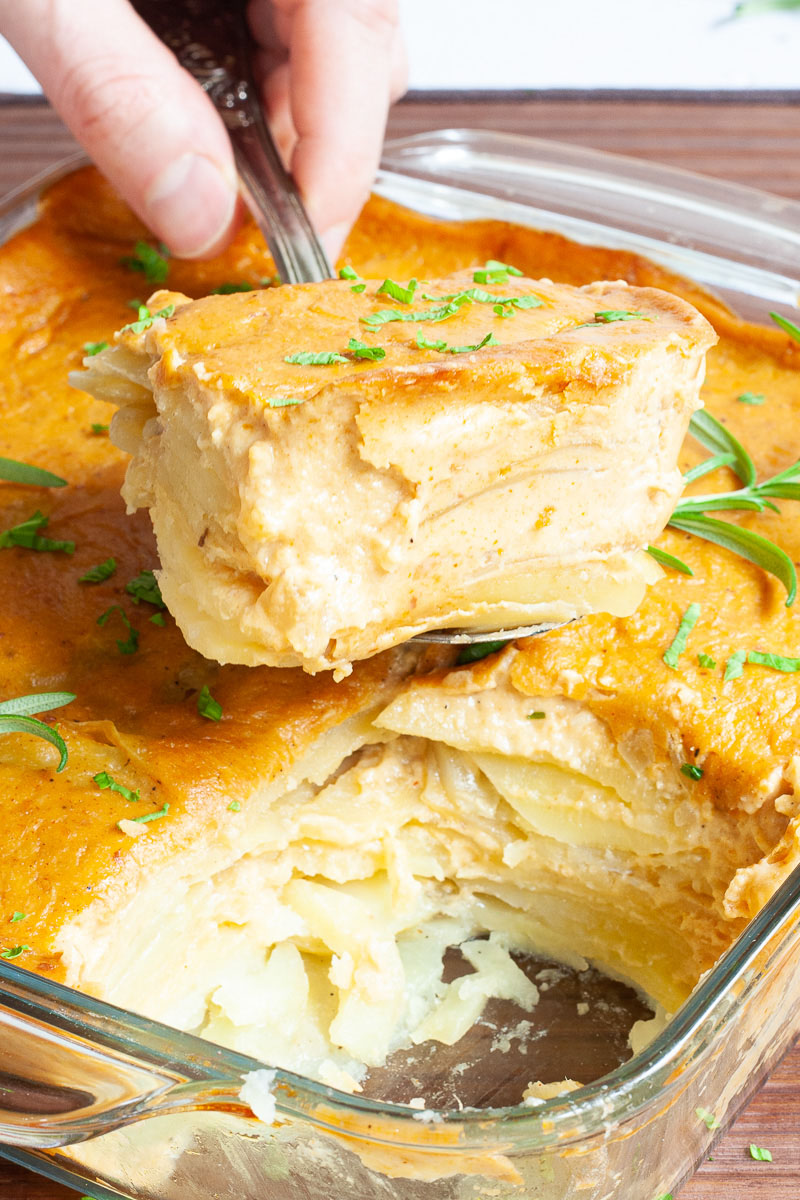 A glass casserole dish with layers of potato slices with an orange creamy sauce sprinkled with chopped parsley. A hand is take a serving size.