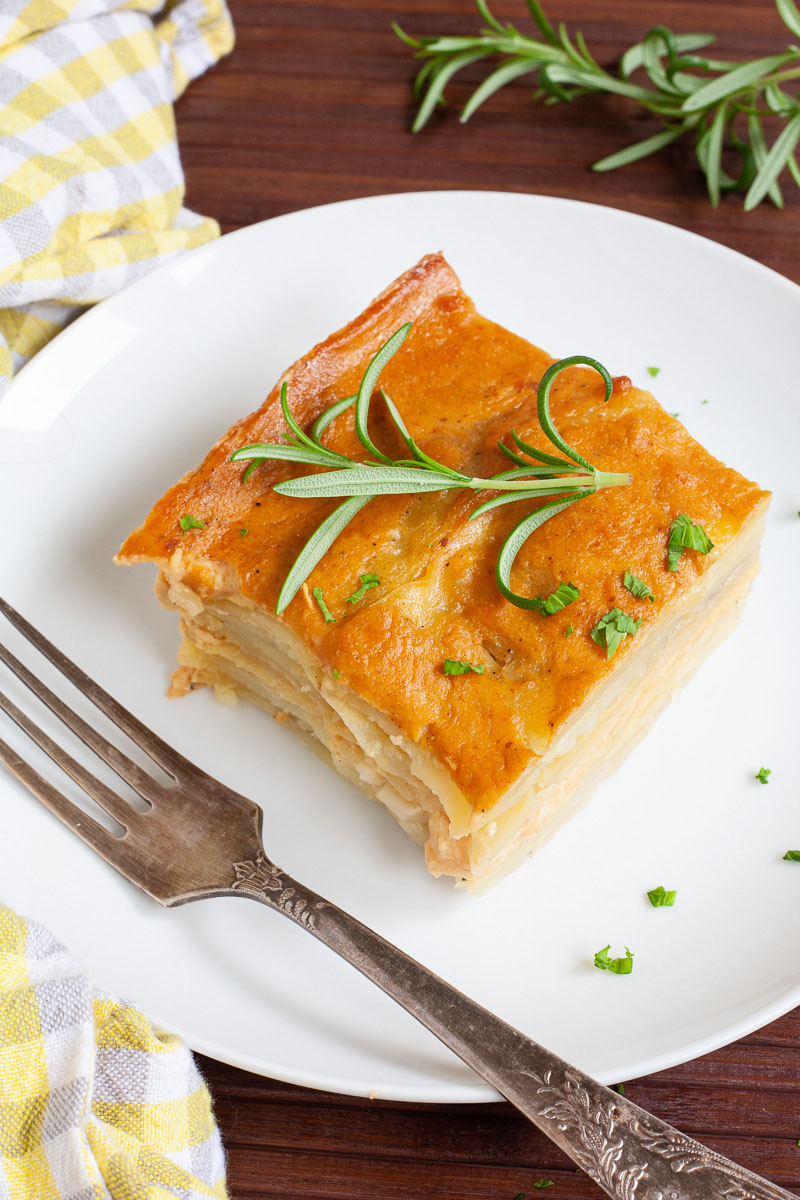 Layers of potato slices with an orange creamy sauce sprinkled with chopped parsley is served on a white plate.