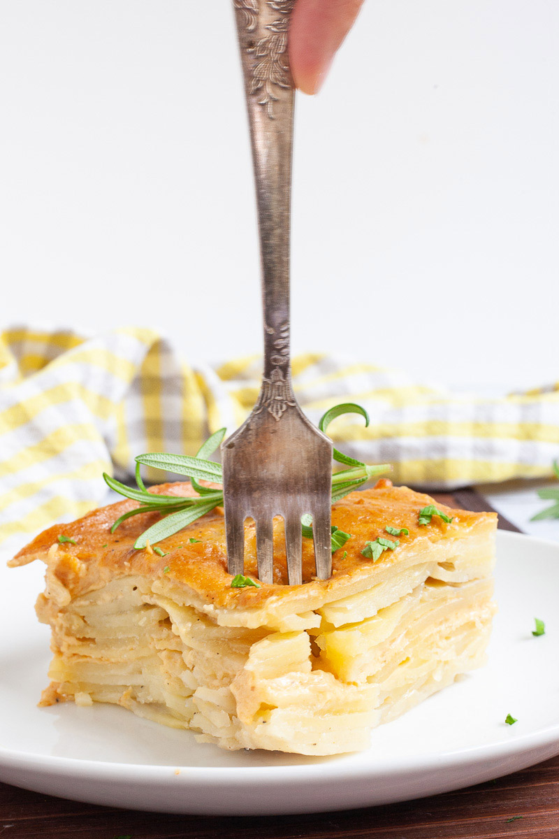 Layers of potato slices with an orange creamy sauce sprinkled with chopped parsley is served on a white plate. A hand is take a bite from above.