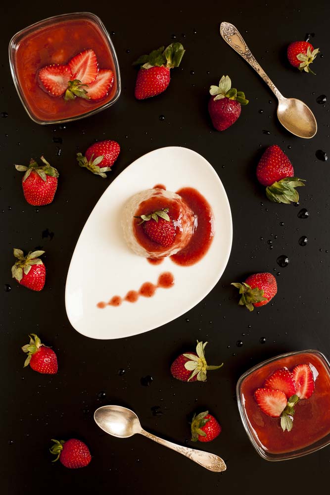 Vegan tapioca pudding topped with strawberry puree and served as panna cotta on a white plate. Black background. Scattered strawberries. 