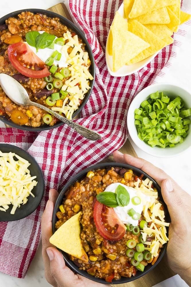 Two hands are holding a black bowl of chili topped with tomato, spring onion, sour cream, grated cheese, basil leaves and nacho chips. Remaining toppings are in small bowls around it. 