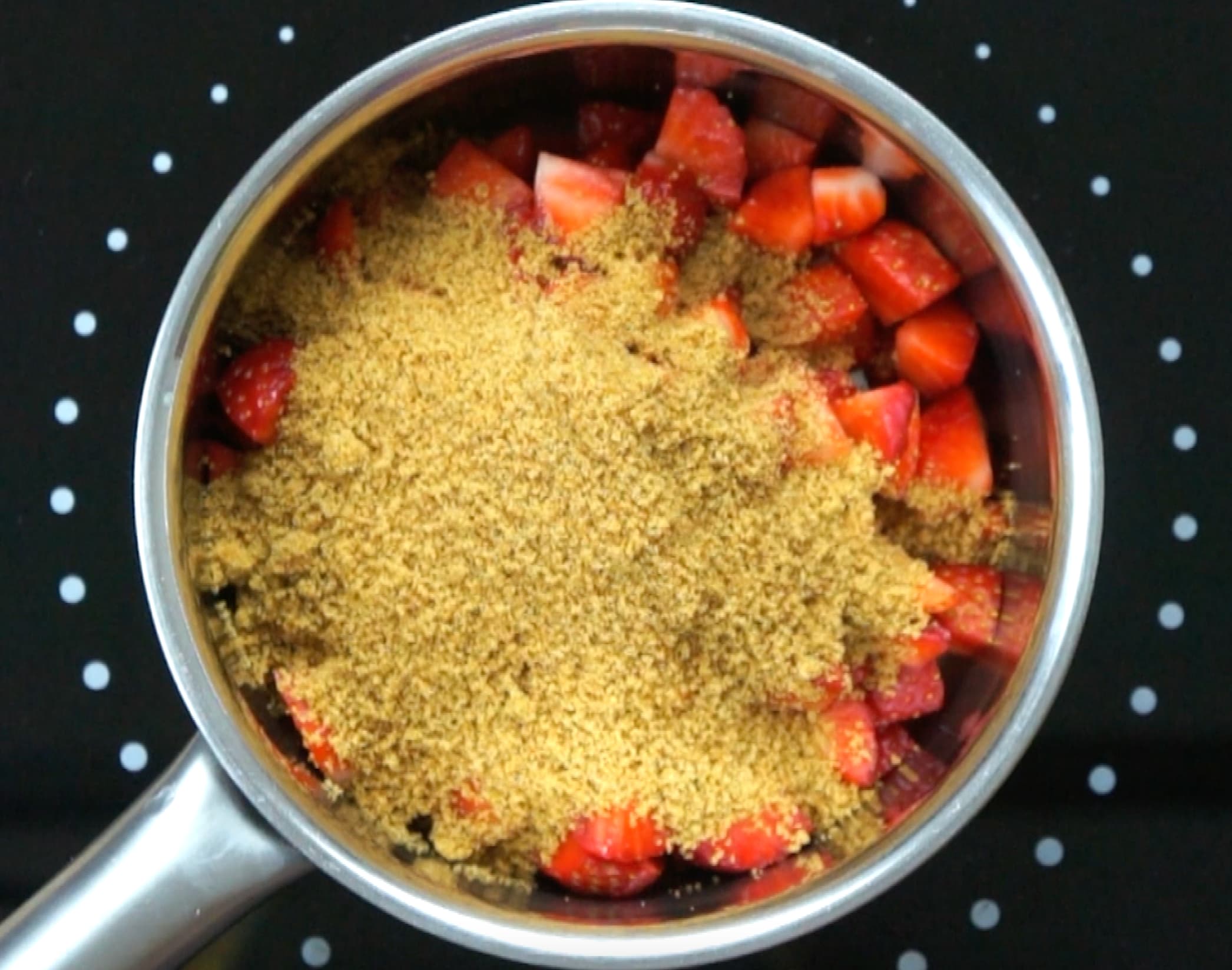 Strawberry pieces in a small saucepan with a heap of brown sugar.